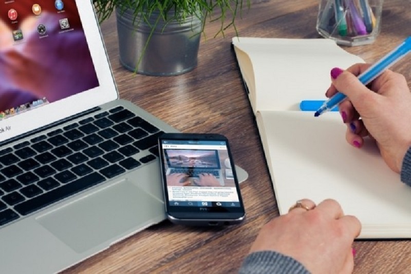 woman-sitting-at-table-writing-in-notepad-with-laptop-and-mobile-phone-on-wooden-table.jpg